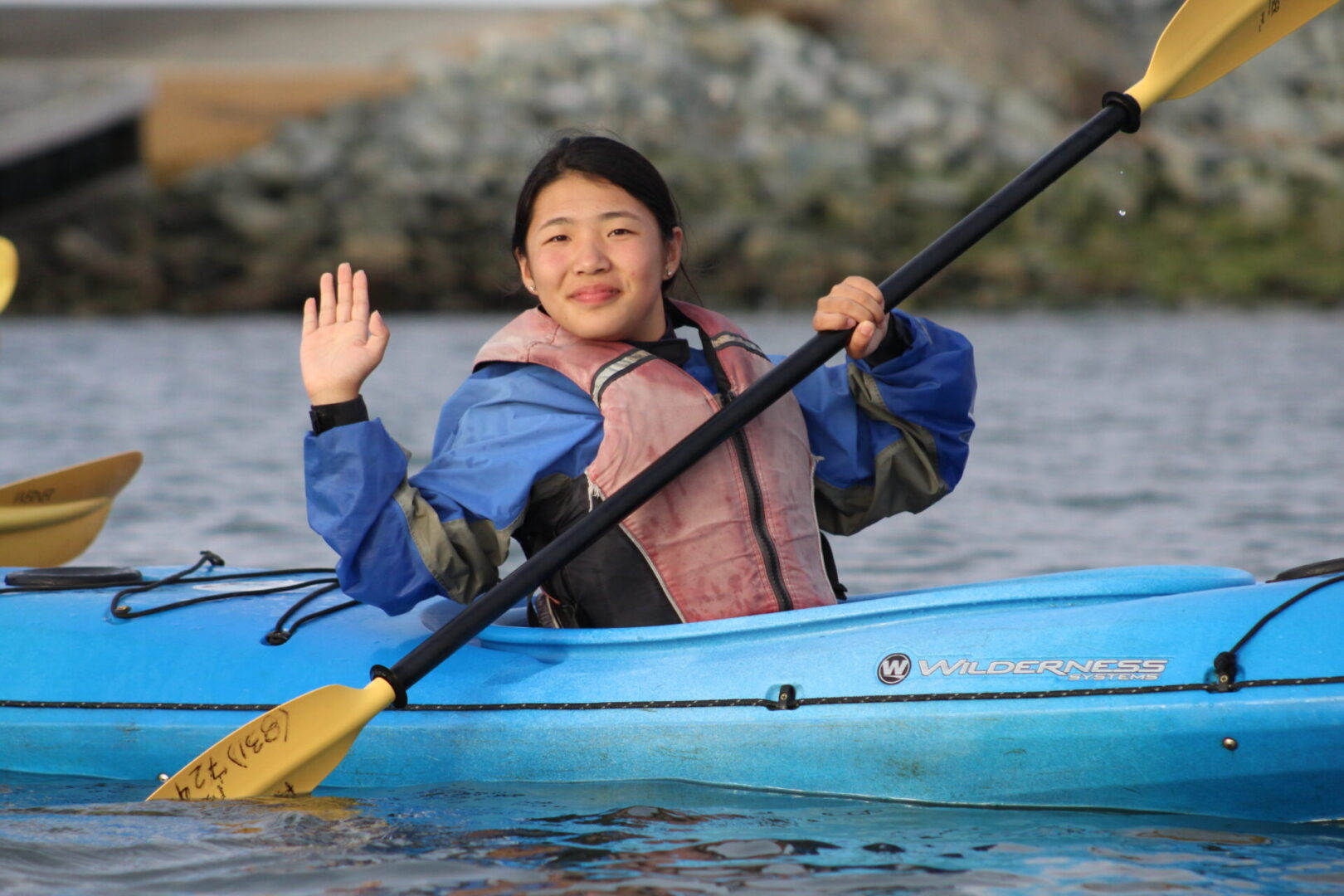 A woman in a blue canoe waving at the camera.