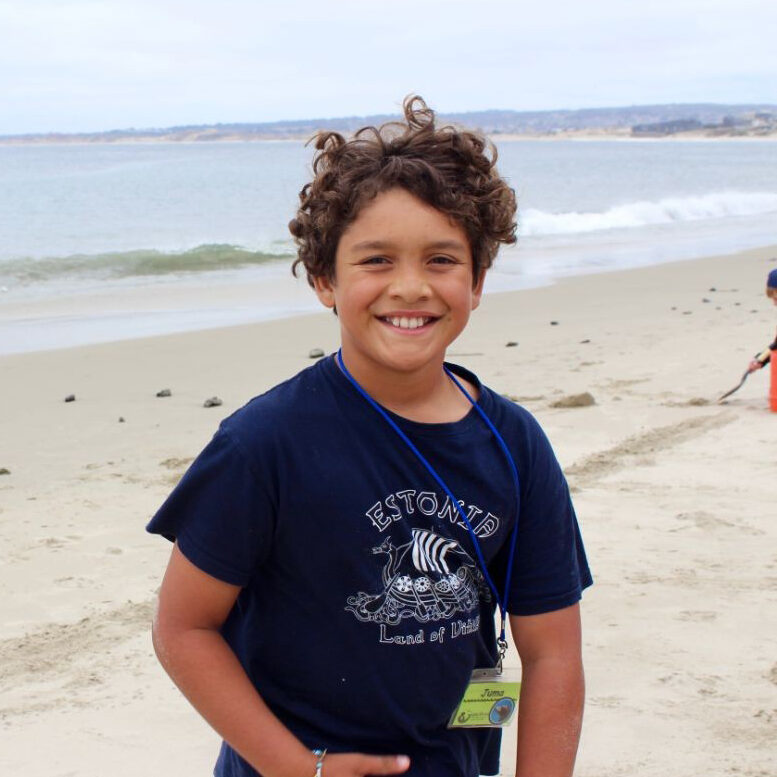 A young boy standing on the beach smiling.